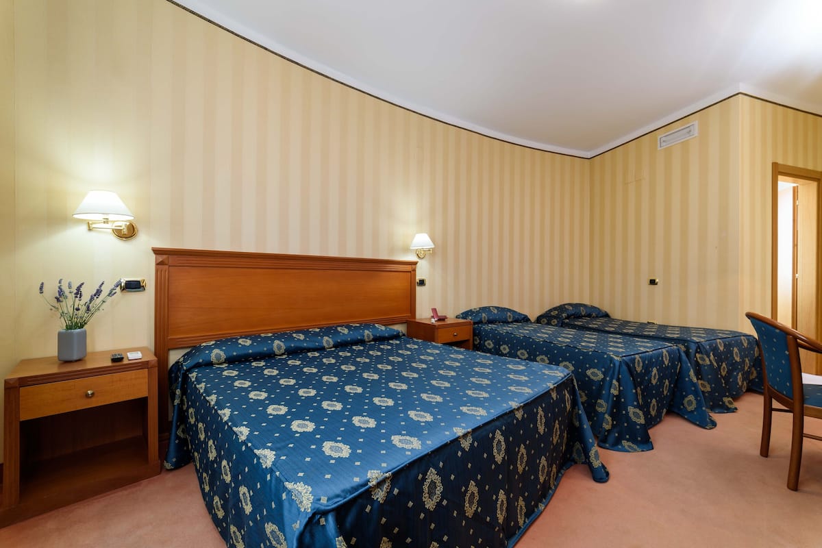A hotel room with two beds, a wooden headboard, bedside tables with lamps, a small desk, and a chair.