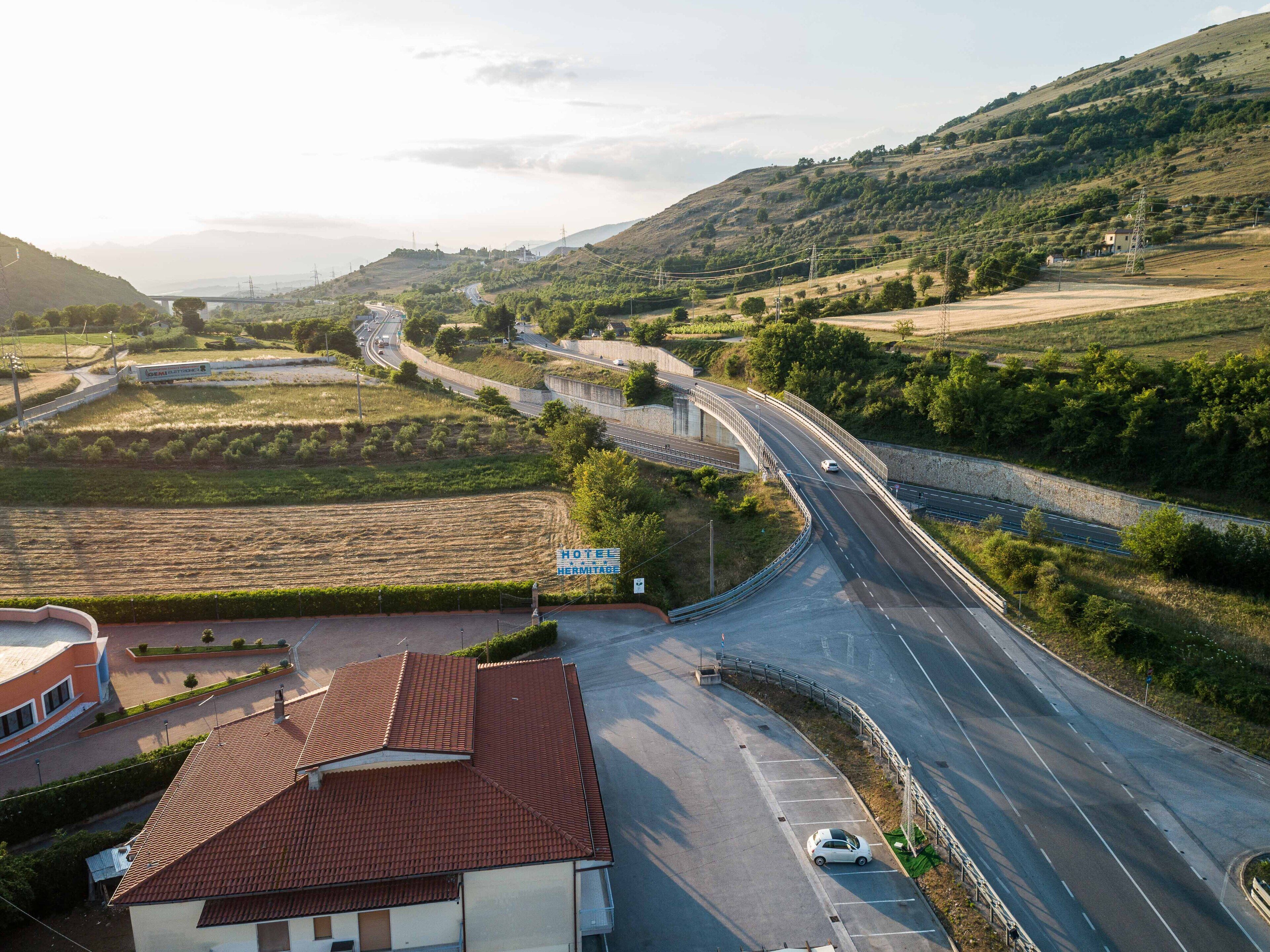 A hotel with a red-tiled roof, a parking lot, and a road curving through a hilly landscape.