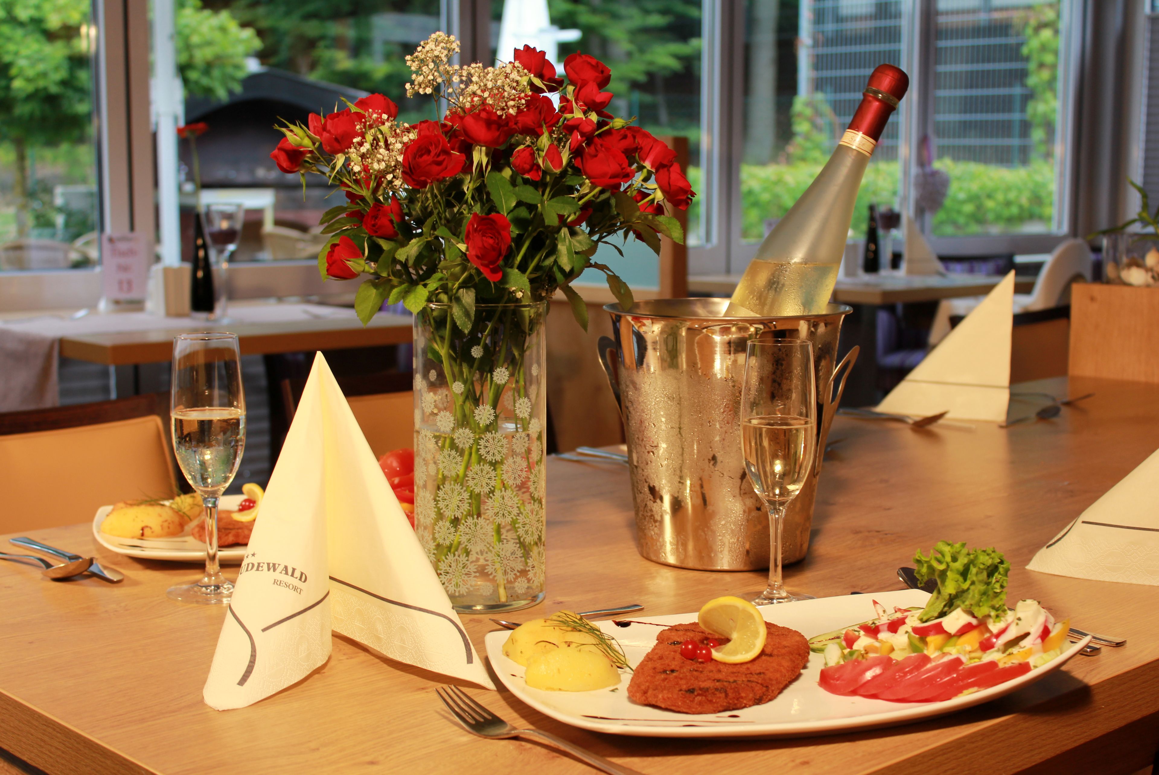 A dining table with a bottle of champagne, a vase of red roses, and a plate of food.