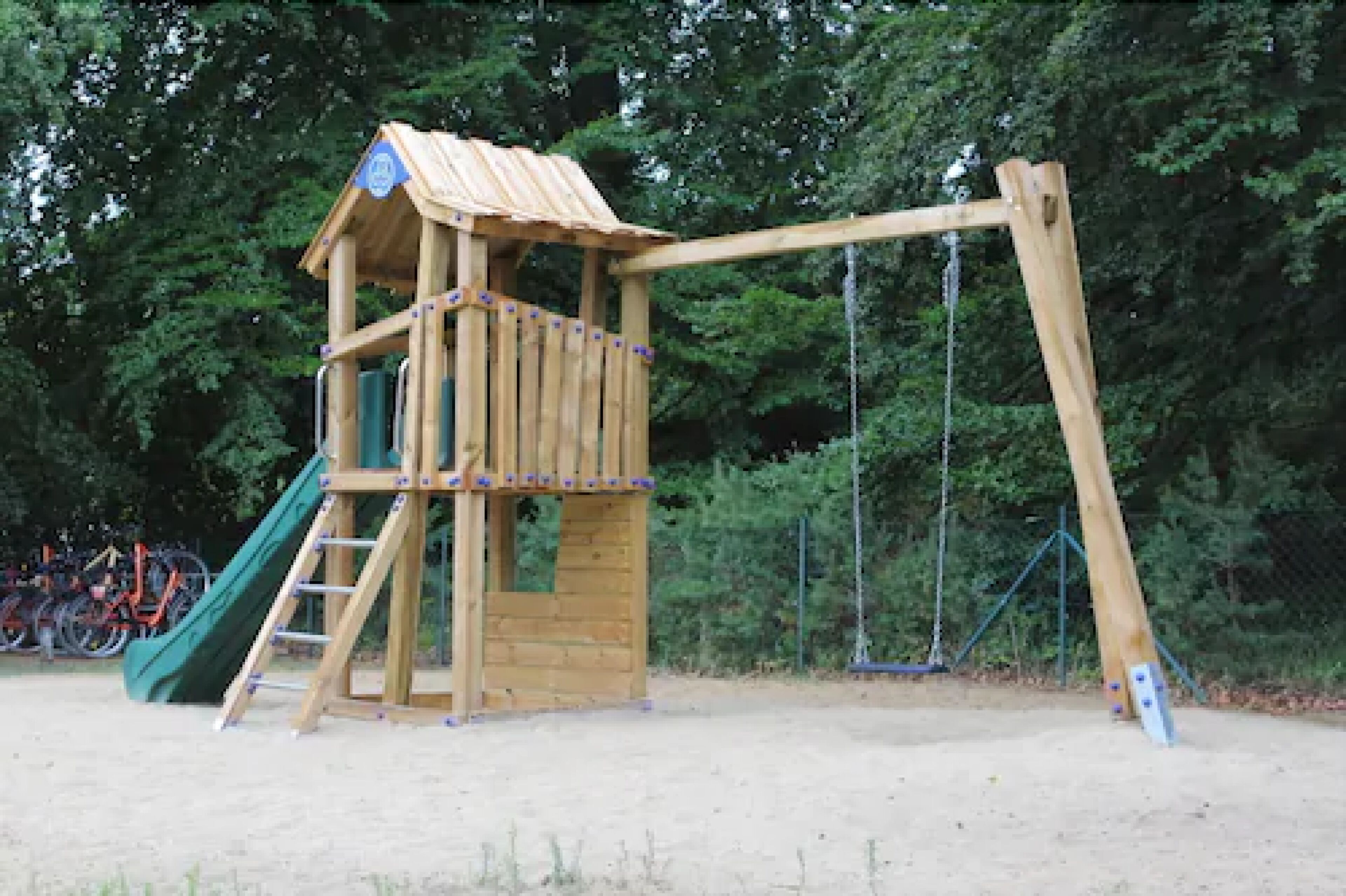 A wooden play structure with a slide and swings, surrounded by trees.