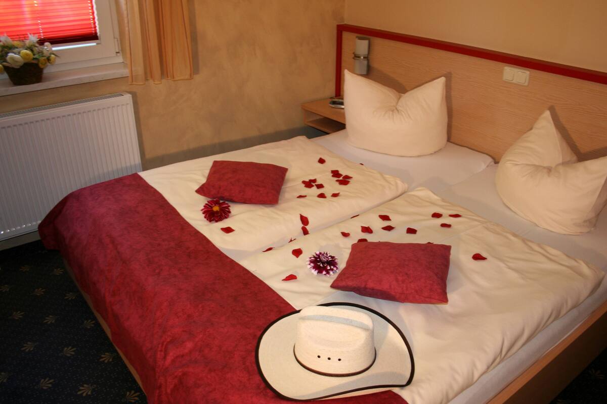 A neatly made bed with red and white bedding, a white hat, and scattered rose petals.