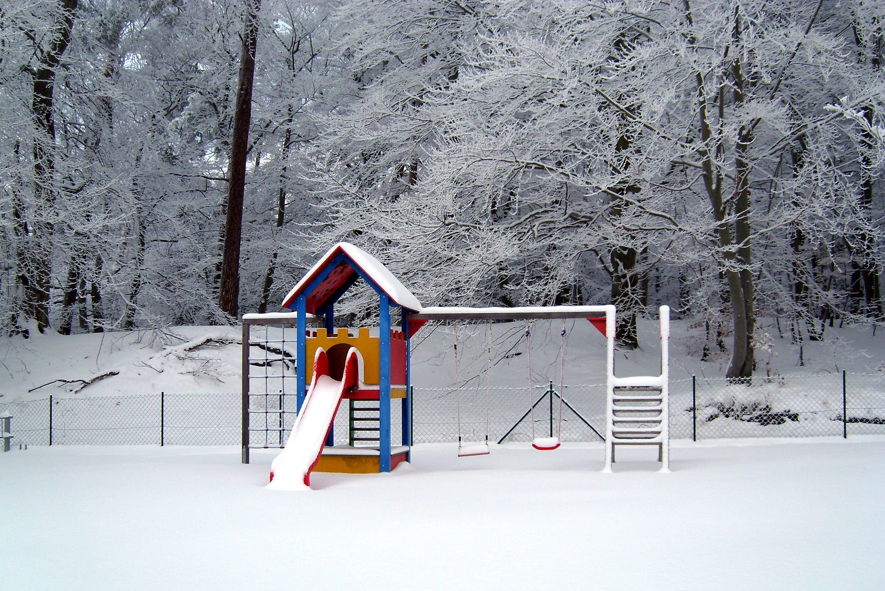 A snow-covered playground with a slide and swings, surrounded by trees.
