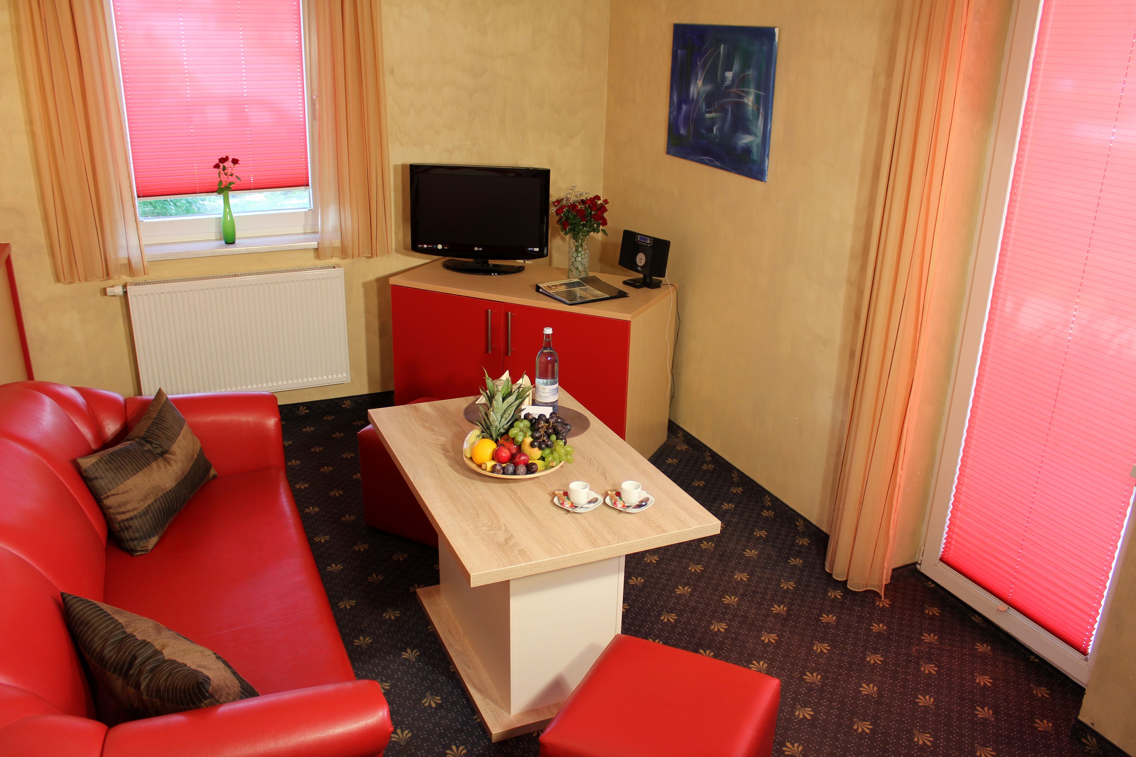 A red sofa, a wooden table with a fruit bowl, a television, and a small cabinet with a vase.