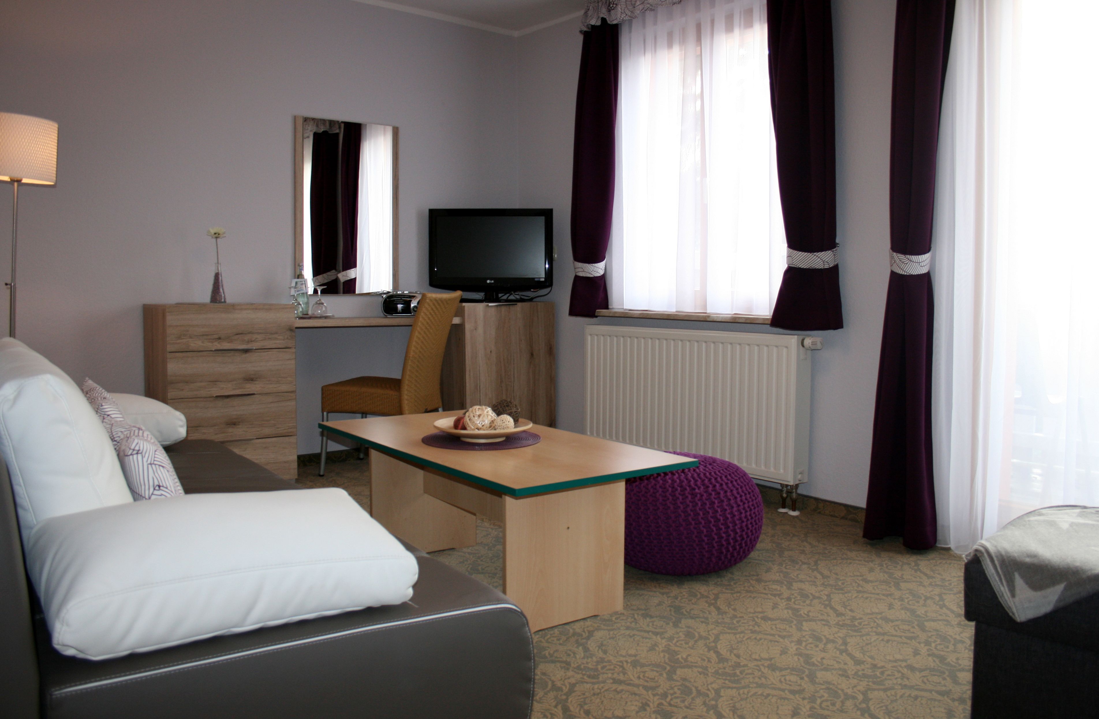 A hotel room with a sofa, a wooden desk, a television, a radiator, and purple curtains.