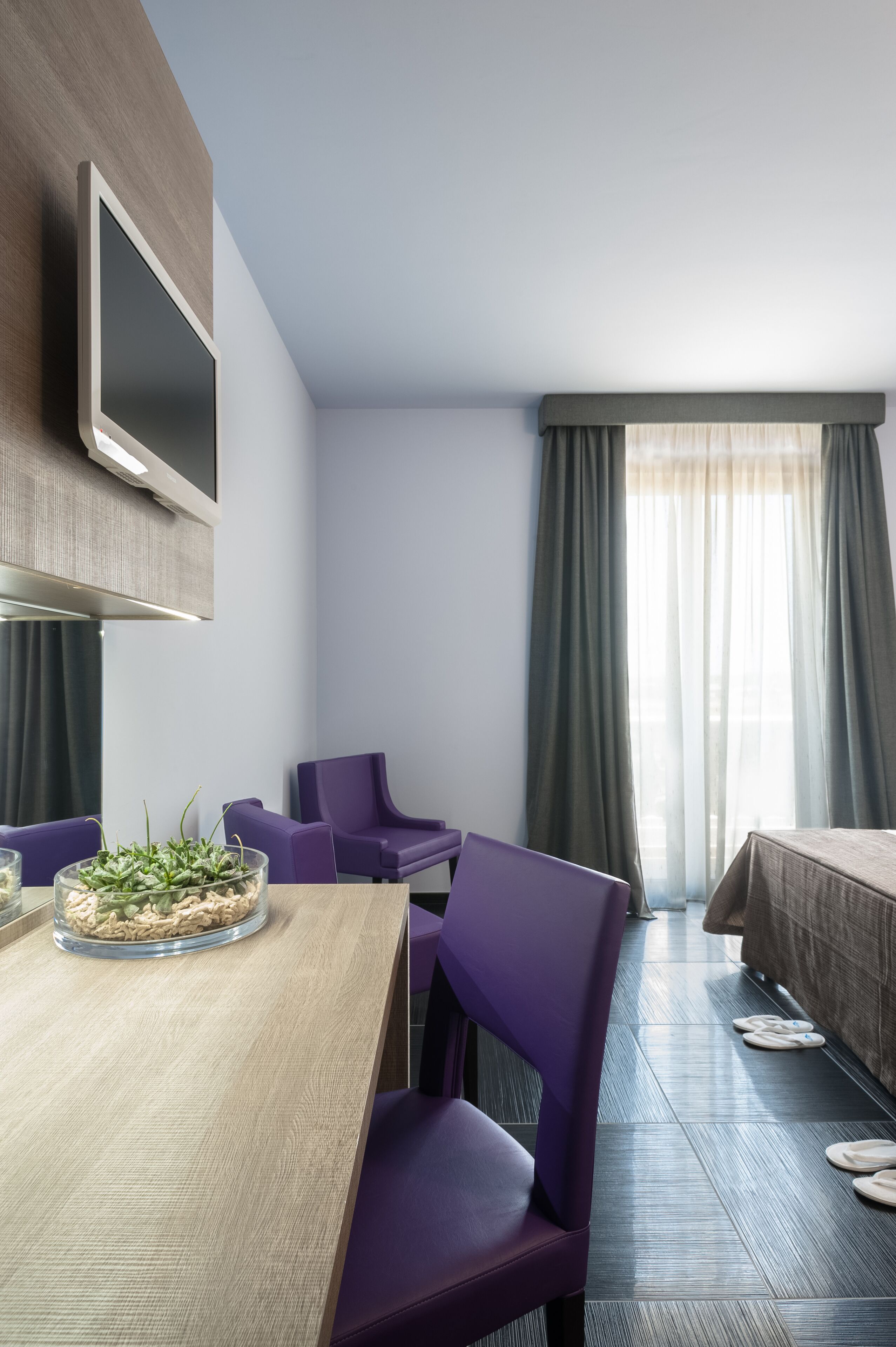 A modern hotel room with a wooden dining table, purple chairs, a flat-screen TV mounted on the wall, a bed with a beige cover, and a window with sheer curtains.