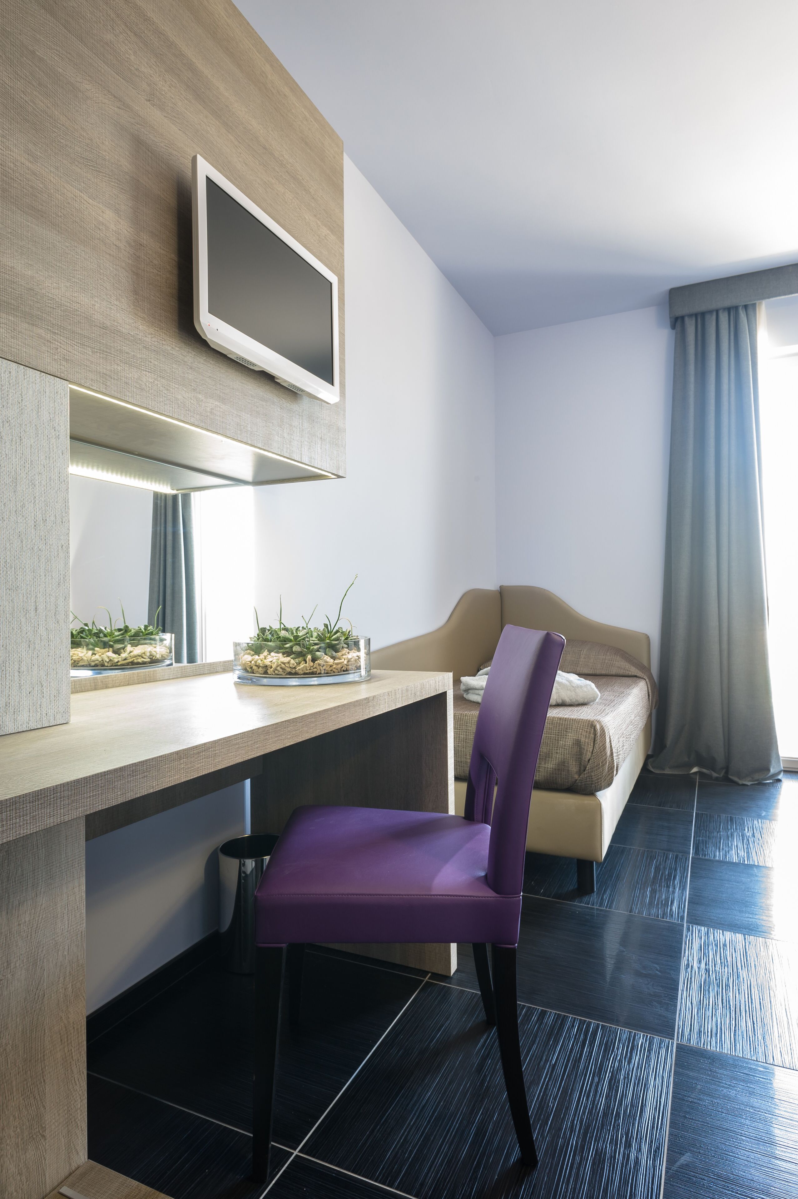 Moderni hotellihuone, jossa on sänky, työpöytä tuolilla, taulutelevisio ja pieni kasvi työpöydällä.