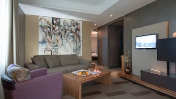 A modern living room with a grey sofa, a wooden coffee table with fruits, a large abstract painting, and a flat-screen TV on a dark wall.
