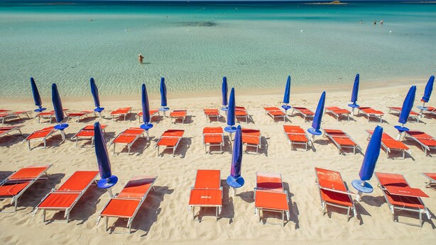 Een strand met rijen oranje ligbedden en blauwe parasols, netjes gerangschikt.