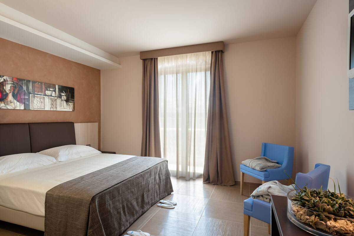 Ein Hotelzimmer mit einem großen Bett, einem Sessel, einem kleinen Tisch und einem Fenster mit Vorhängen.
