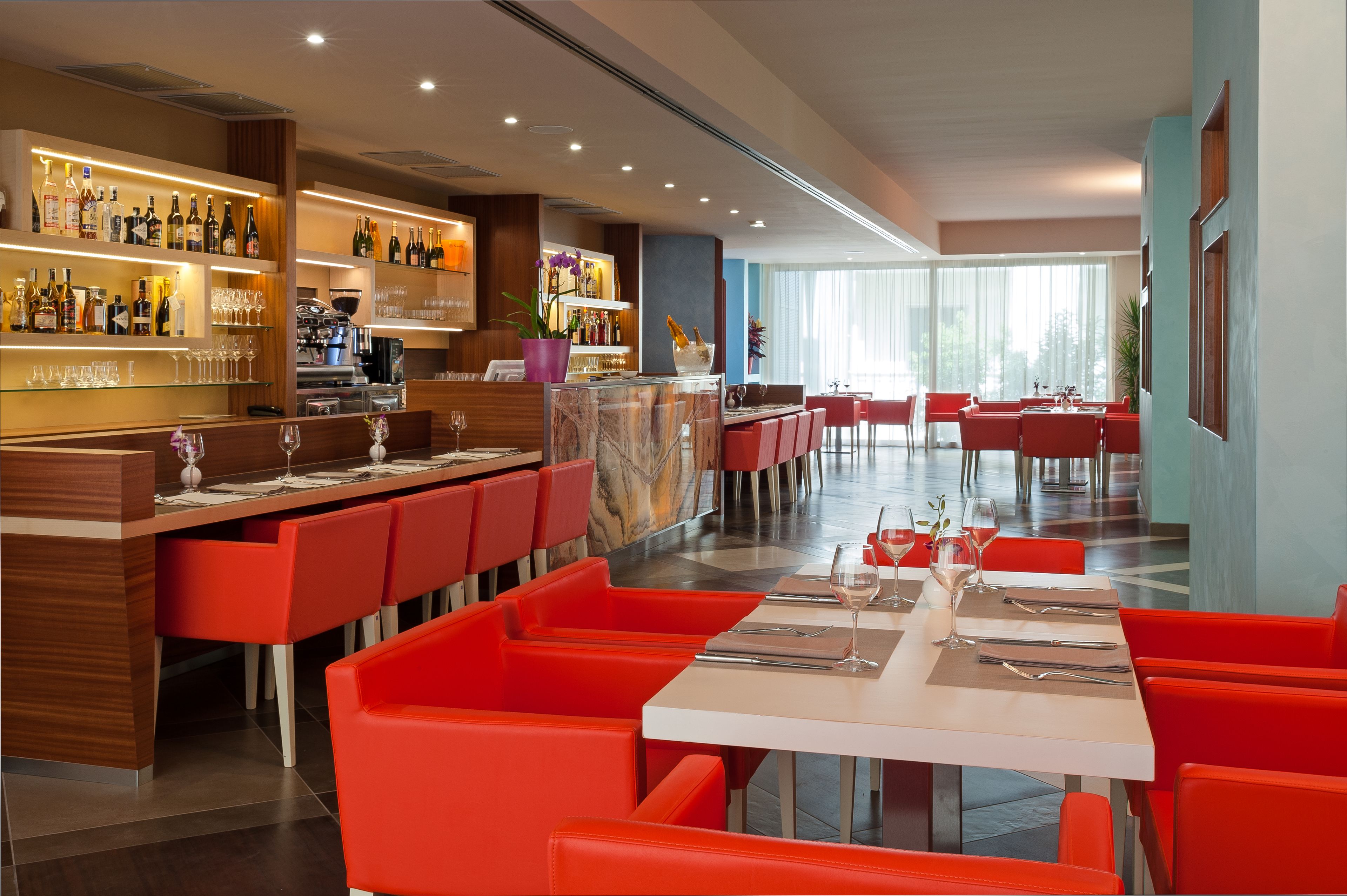 Un restaurant moderne avec des chaises rouges et un bar où l’on peut voir diverses bouteilles et différents types de verres.