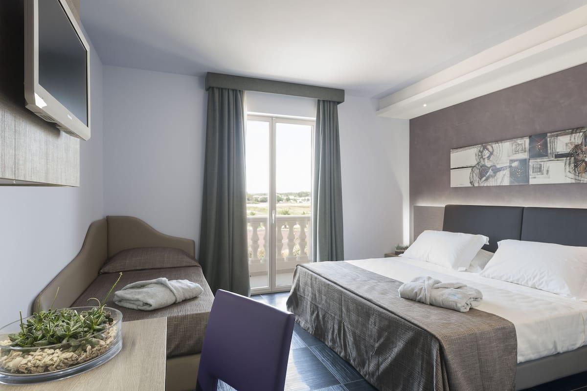 Ein Hotelzimmer mit Bett, Sessel, kleinem Tisch mit Pflanze, Fernseher und Balkon mit Vorhängen.