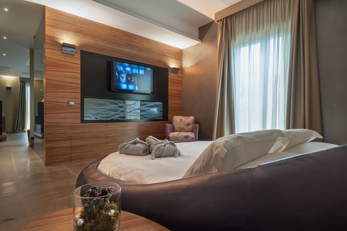 Ein modernes Hotelzimmer mit einem großen Bett, einem flachbildfernseher an der Wand, einer Sitzecke mit einer gepolsterten Bank und einer dekorativen Vase auf dem Nachttisch.