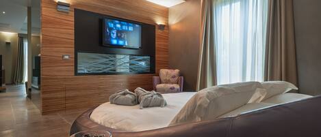 A modern hotel room with a large bed, a flat-screen TV mounted on the wall, a seating area with a cushioned bench, and a decorative vase on the side table.