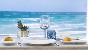 Een gedekte tafel met borden, glazen en een glas water, met uitzicht op een strand met golven en een heldere lucht.