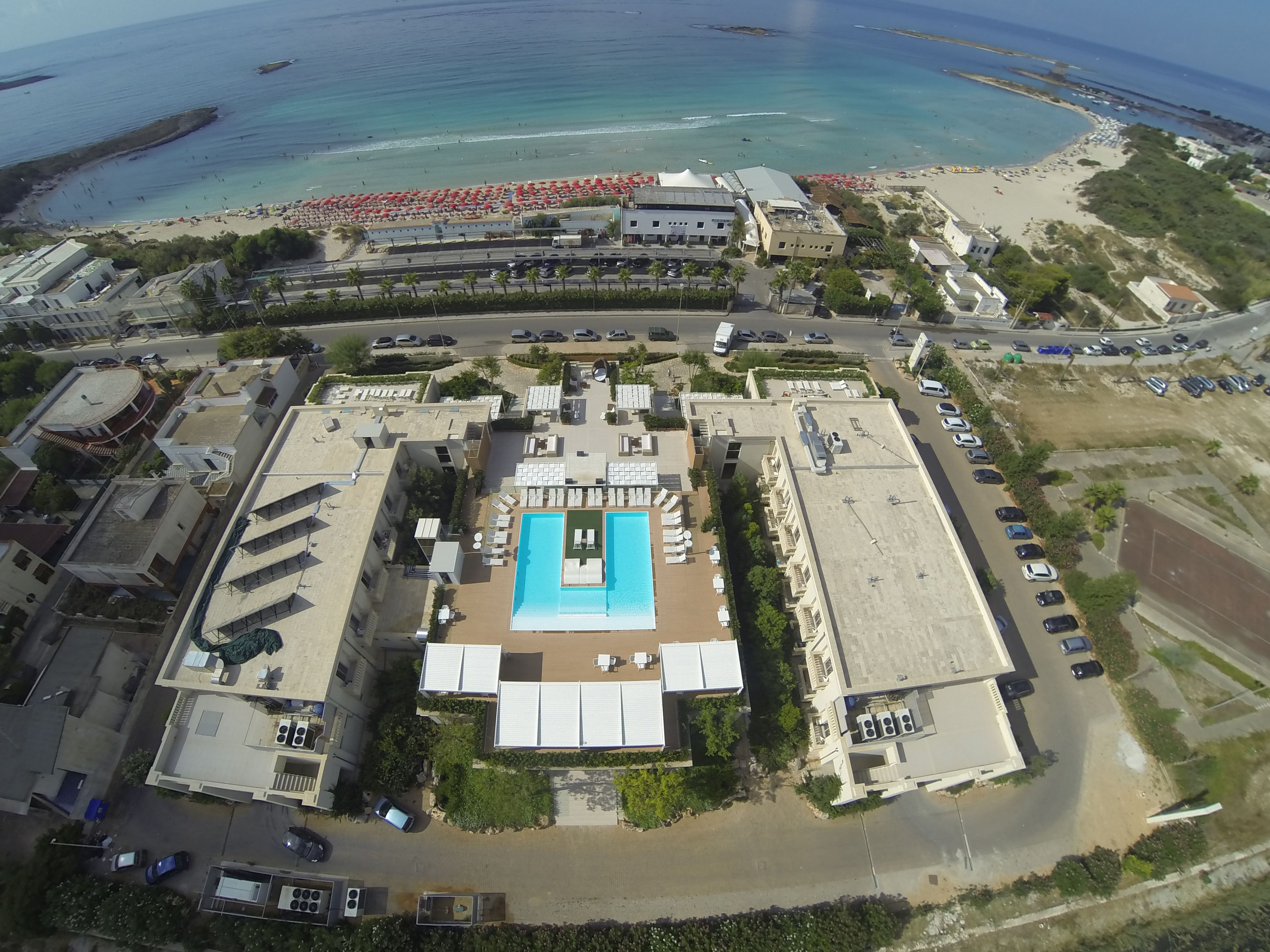 Ovenfra et resort med en central swimmingpool, flere bygninger og et strandområde med parasoller.