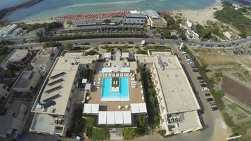 An aerial view of a resort with a central swimming pool, multiple buildings, and a beach area with umbrellas.