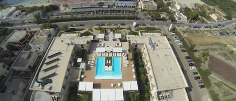 An aerial view of a resort with a central swimming pool, multiple buildings, and a beach area with umbrellas.
