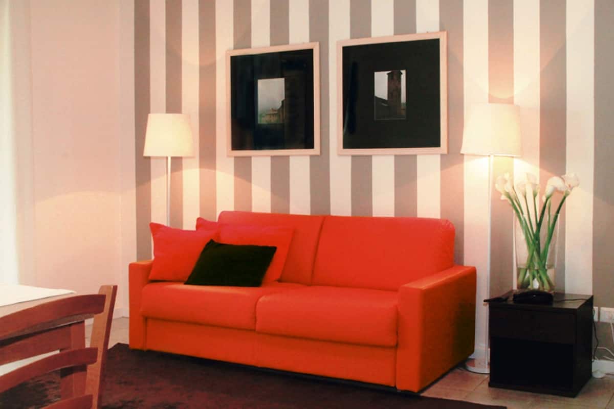 Een kamer met een rode bank, twee ingelijste foto's aan de muur, een lamp en een vaas met bloemen.