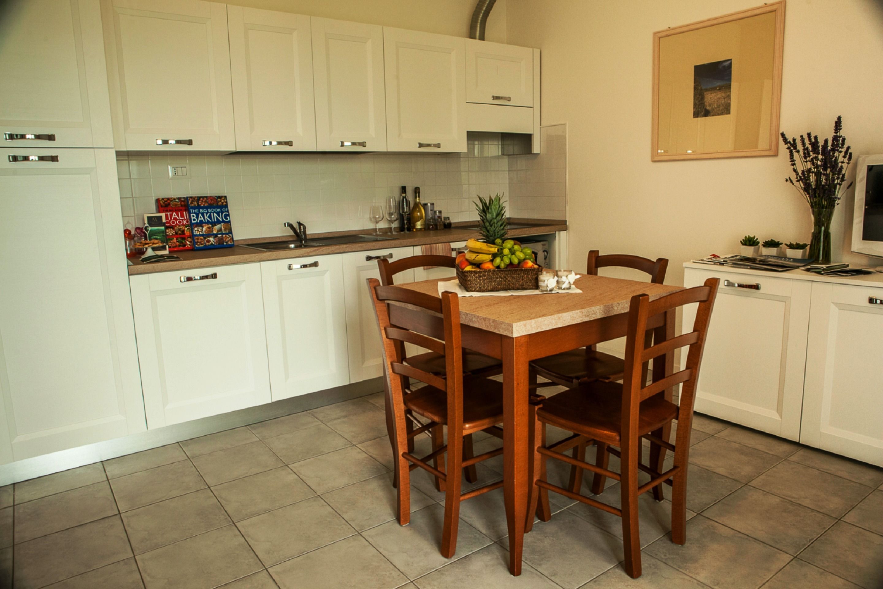 A kitchen with white cabinets, a wooden dining table with chairs, a fruit basket, and a vase with flowers.