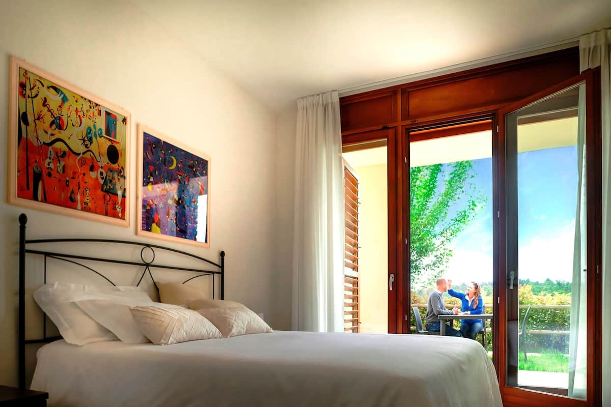 Een slaapkamer met een bed, twee schilderijen, een raam met uitzicht op een wijngaard en een schuifdeur naar een buitengebied.