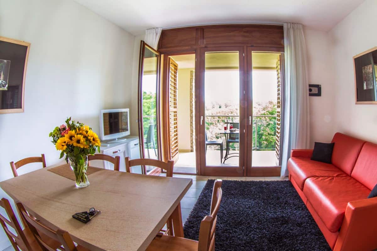 Een kamer met een eettafel, stoelen, een rode bank, een televisie en een balkon met uitzicht.