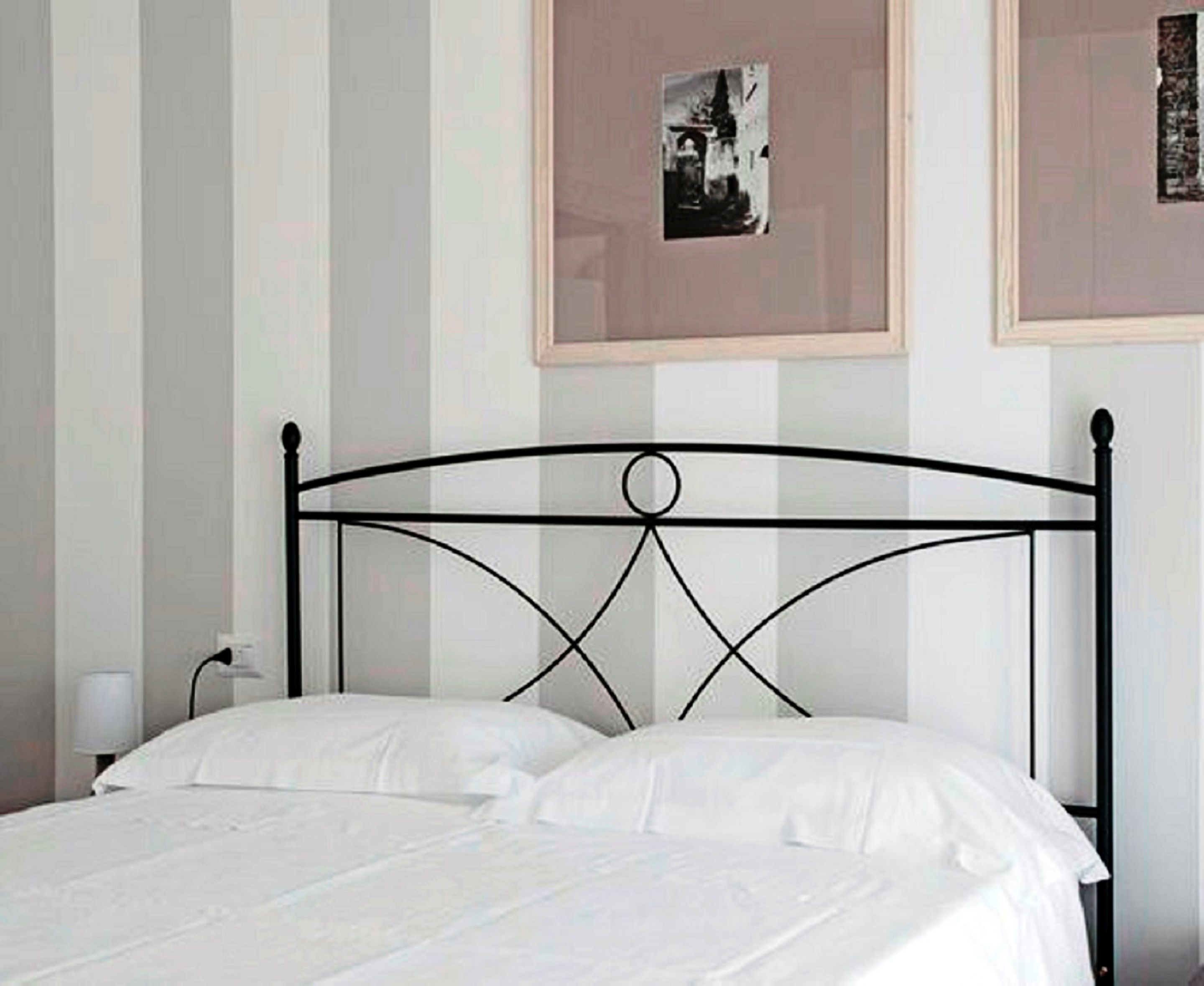 Ein Bett mit Metallgestell und weißer Bettwäsche in einem Zimmer mit Streifenmuster an den Wänden und zwei gerahmten Bildern.