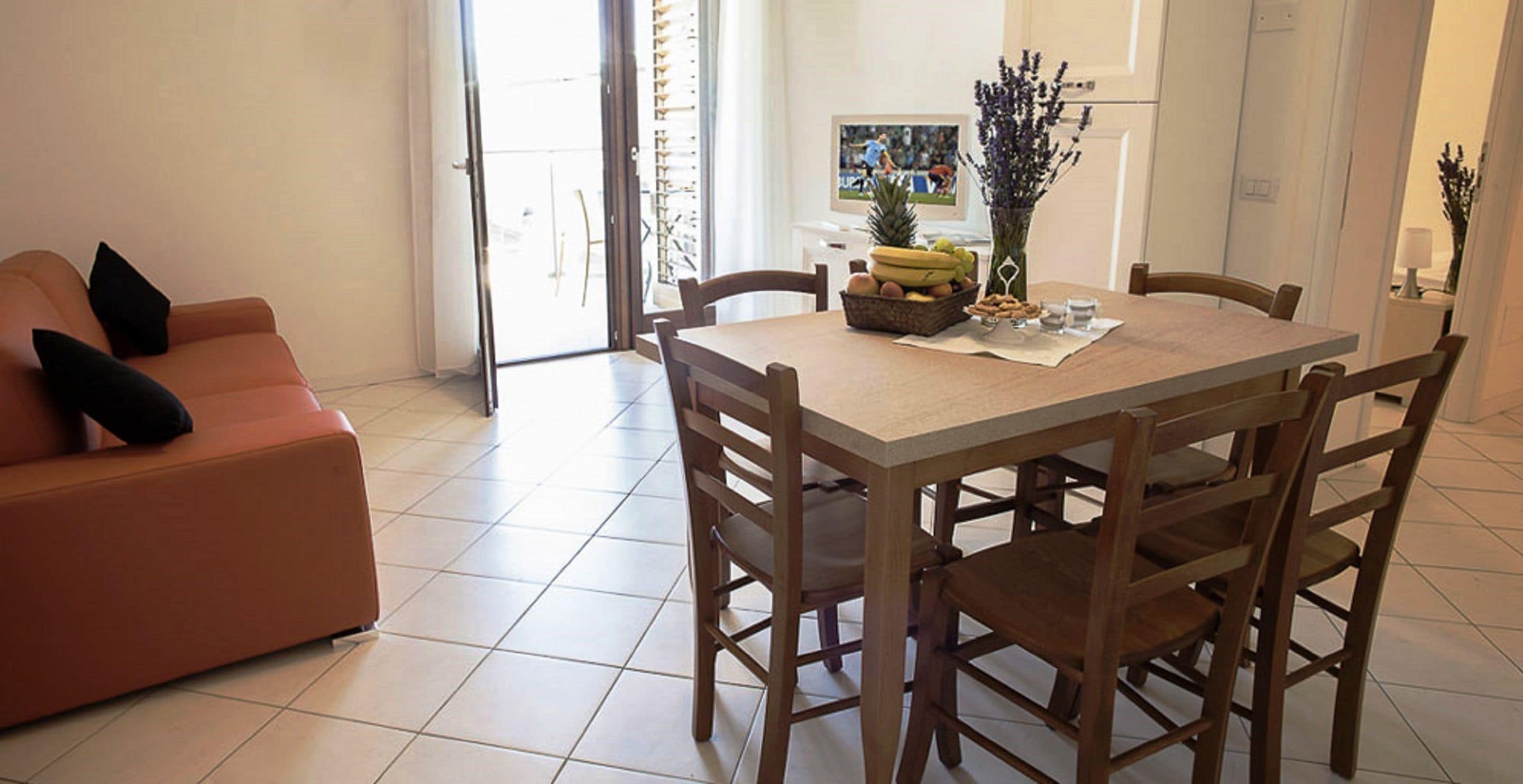 Uma área de refeições com uma mesa posta para quatro pessoas, um sofá com almofadas e uma televisão ao fundo.