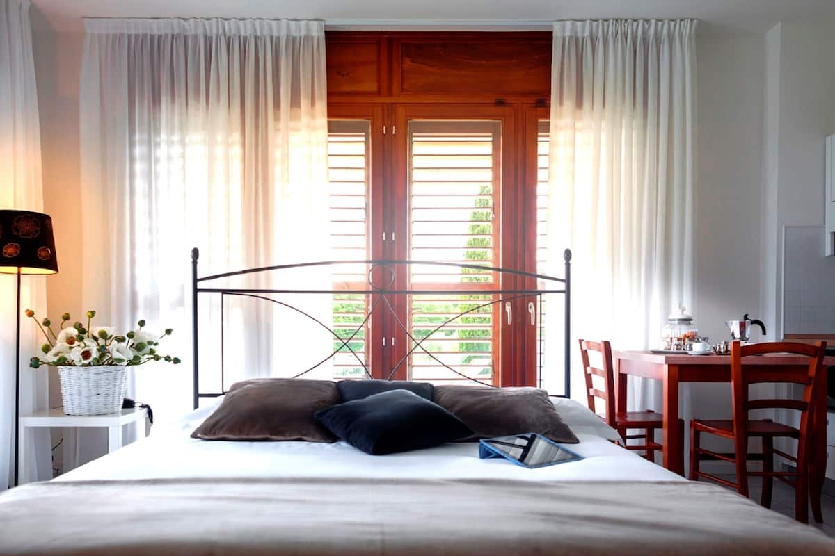 Een slaapkamer met een bed, een nachtkastje met een lamp, een stoel en uitzicht op een balkon met houten luiken.