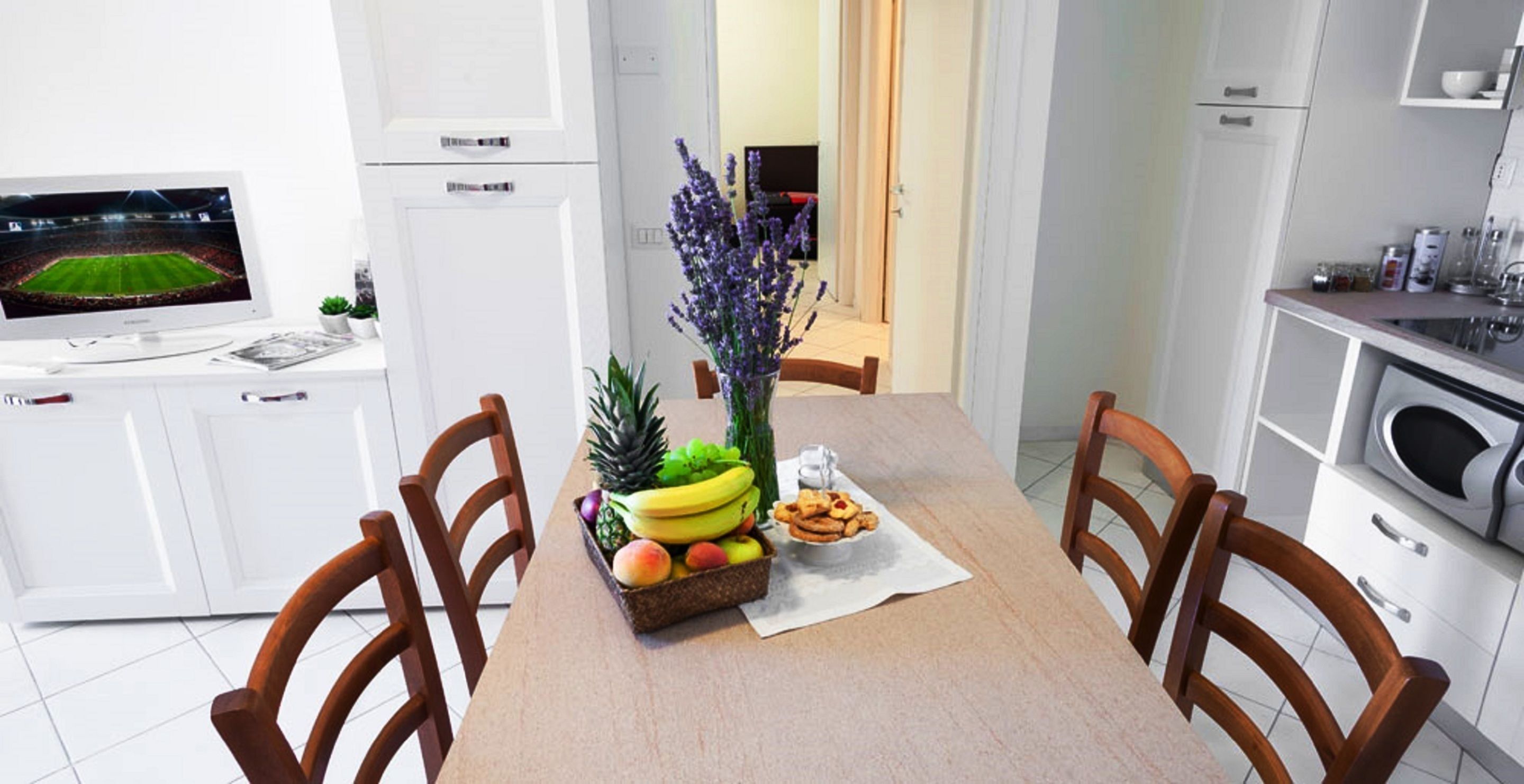 A modern kitchen with white cabinets, a dining table with chairs, a fruit basket, and a vase with flowers.