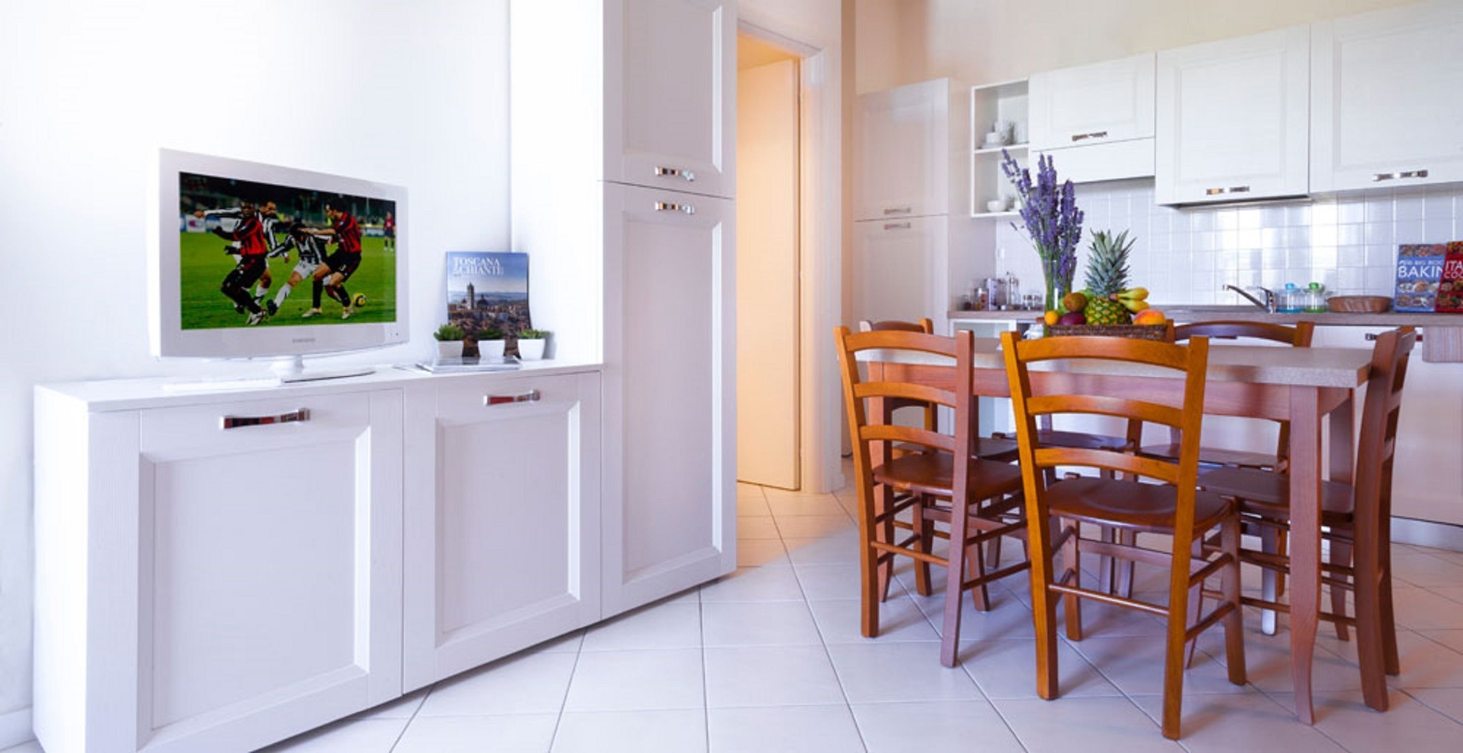 Eine moderne Küche mit weißen Schränken, einem Esstisch mit Holzstühlen und einem Flachbildfernseher auf einem weißen Schrank.