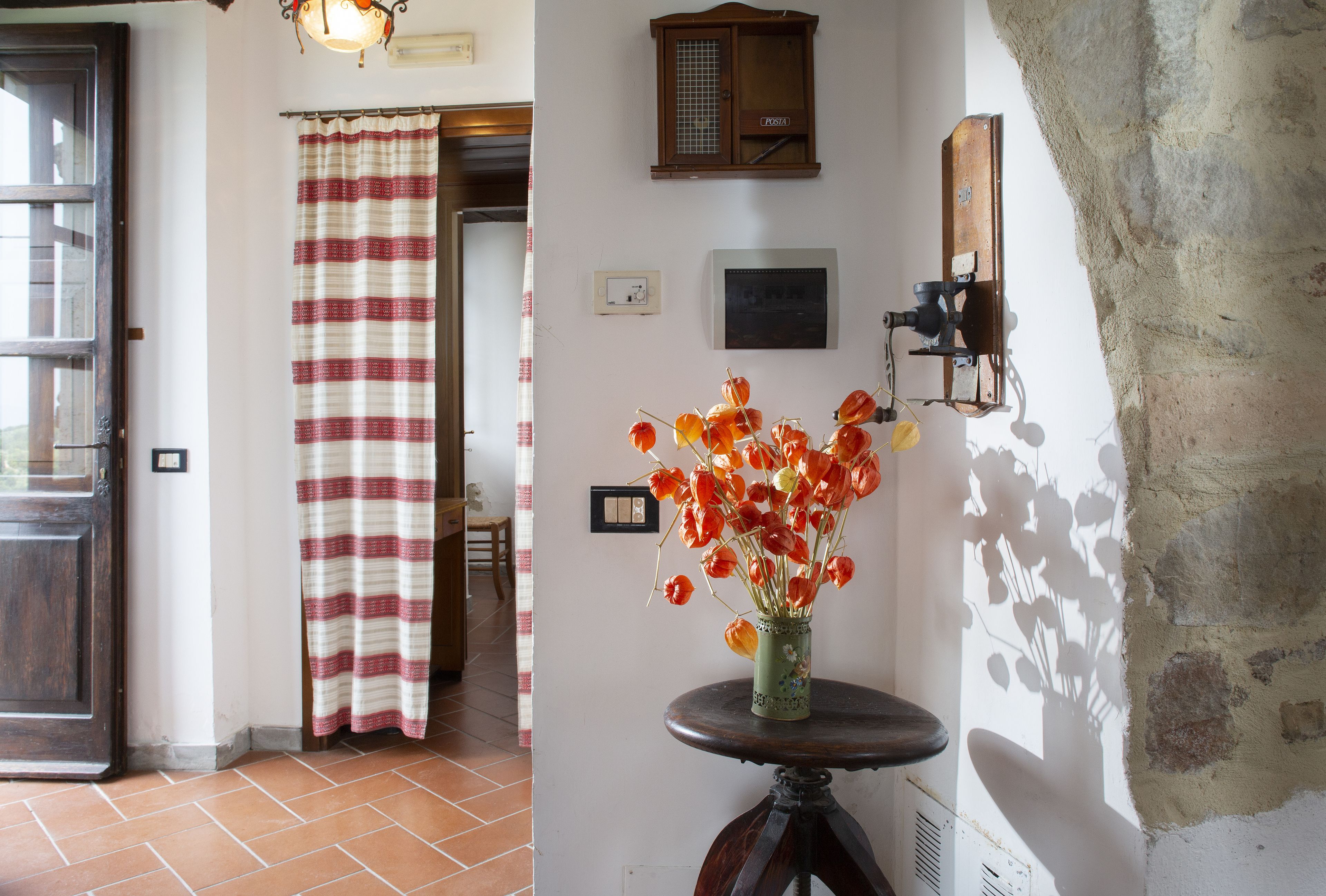 A room with a tiled floor, a wooden door, a striped curtain, a vase with orange flowers, a wall-mounted light fixture, and a small framed picture.