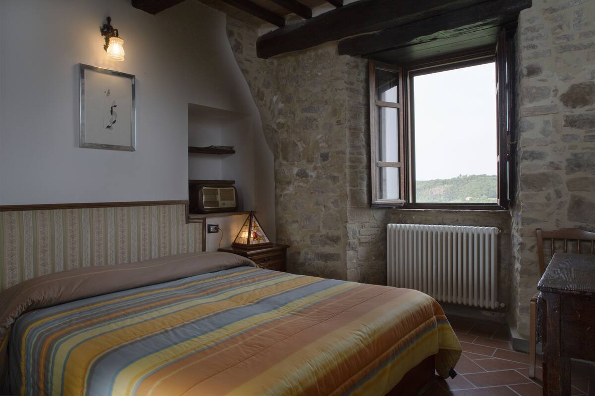 A bedroom with a bed, a nightstand, a lamp, a window with a view, and stone walls.