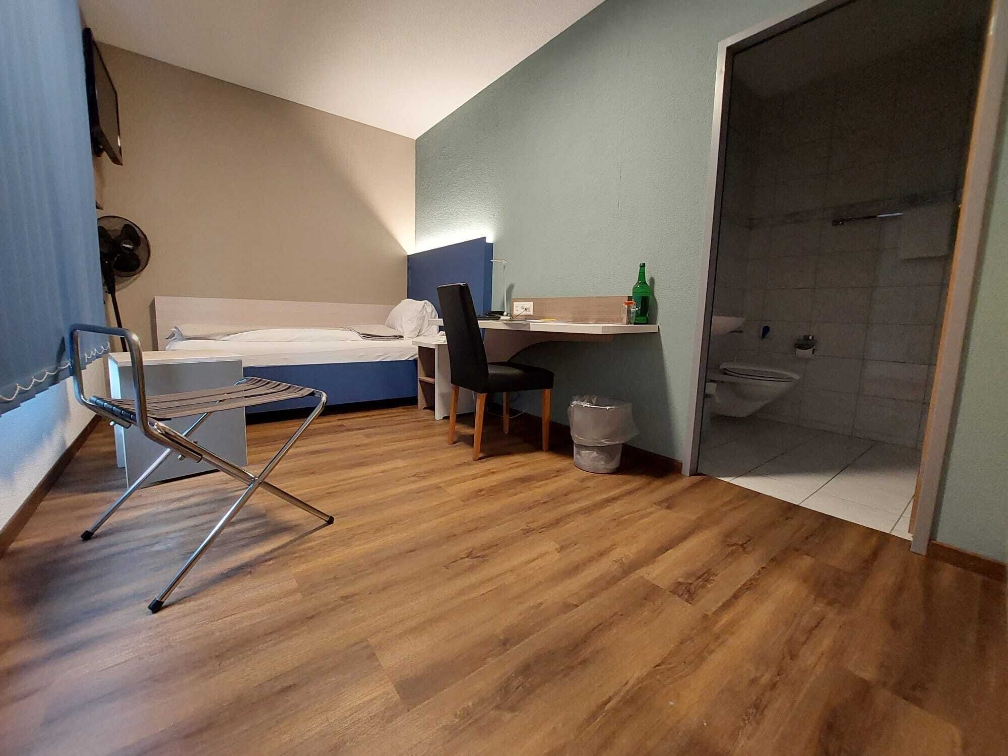 A compact hotel room with a bed, a desk, a chair, a small trash bin, and a shower area.
