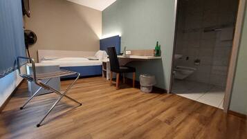 A compact hotel room with a bed, a desk, a chair, a small trash bin, and a shower area.