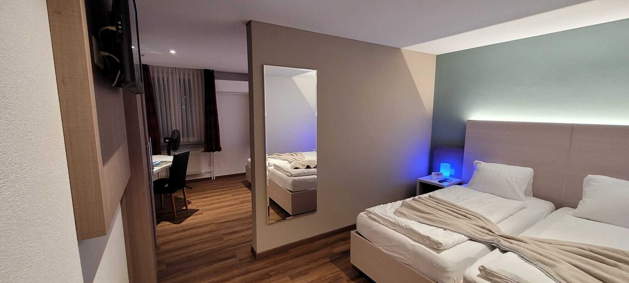 A hotel room with a bed, bedside table with a lamp, a mirror, a desk with a chair, and a window with curtains.
