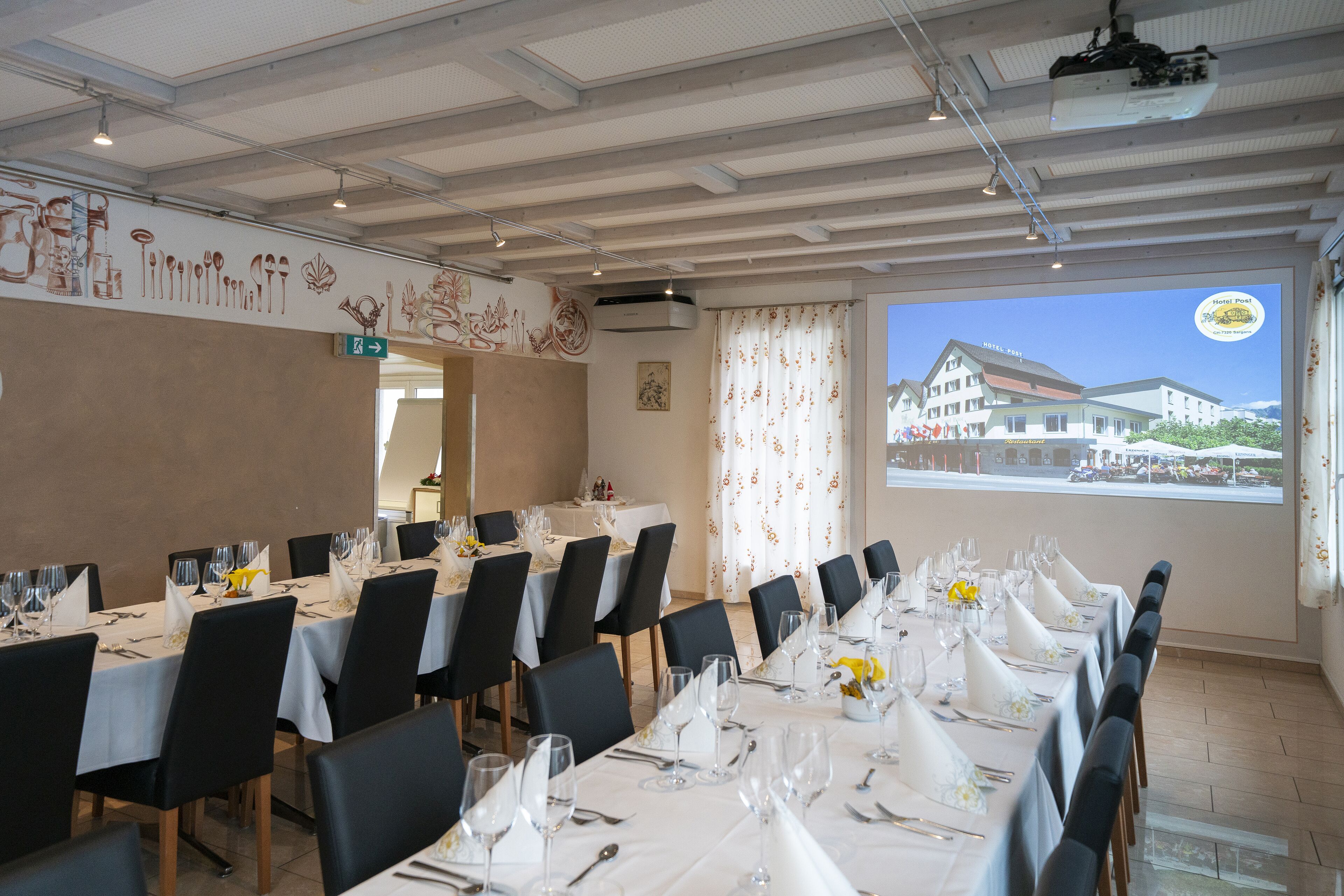 Un salón de conferencias con mesas dispuestas para una reunión, una pantalla de proyector mostrando una imagen de hotel y vistas del edificio del hotel a través de grandes ventanales.