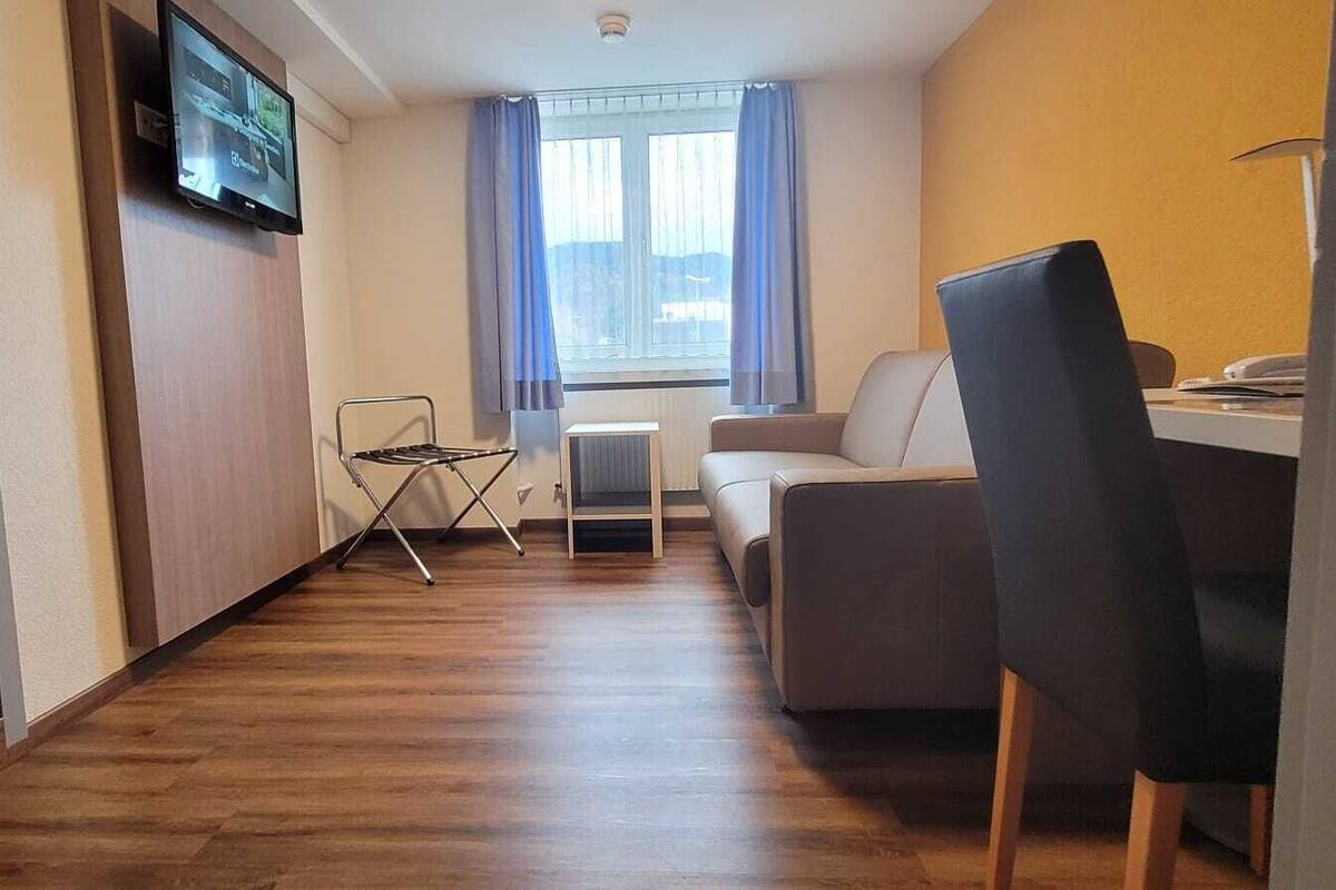 A modern, compact living space with a wooden floor, a flat-screen TV mounted on the wall, a small folding chair, a sofa, and a dining table with chairs.