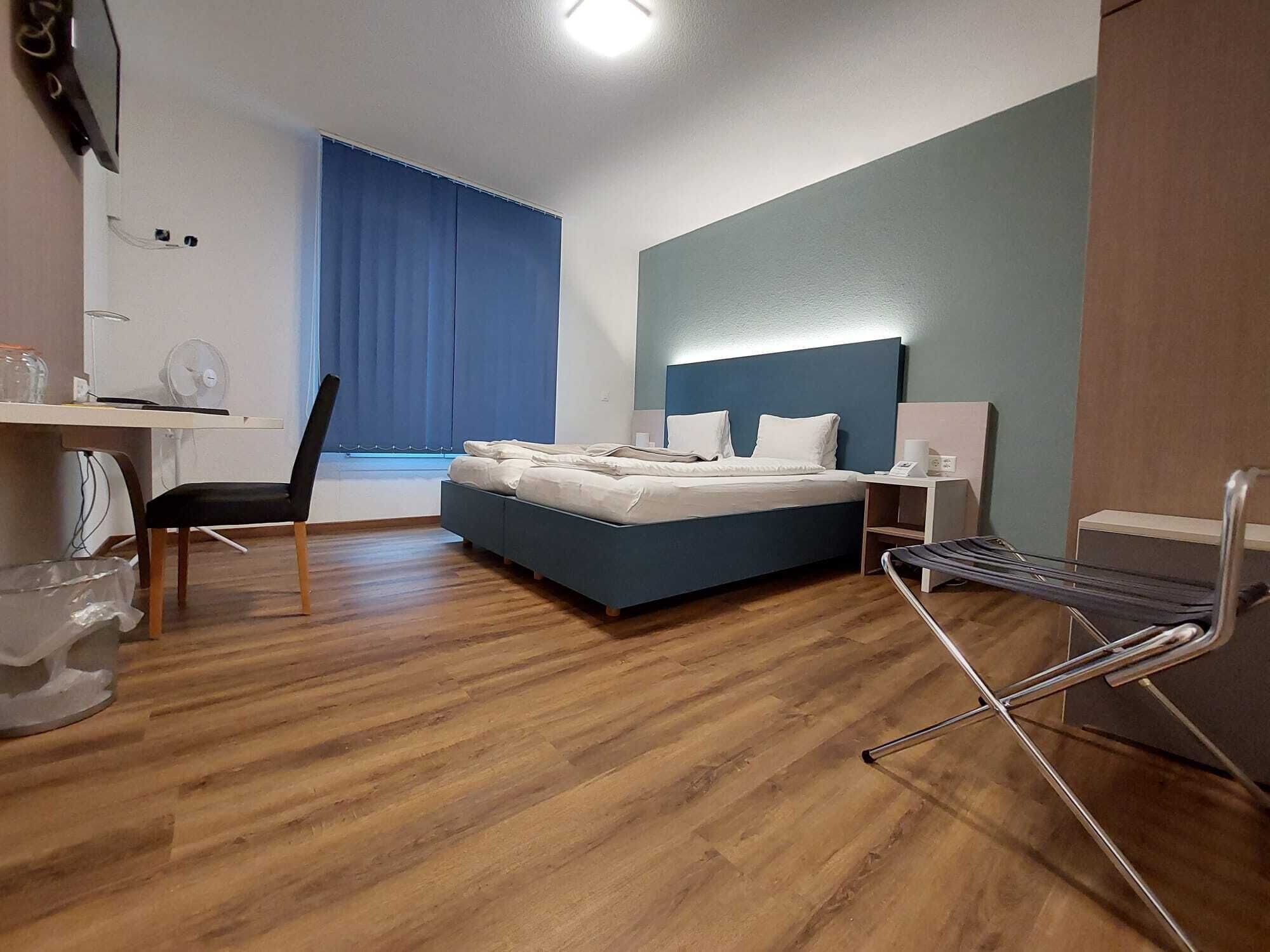 Ein Hotelzimmer mit einem Bett, einem Schreibtisch mit Stuhl, einer Schreibtischlampe und einem Fenster mit Vorhängen.