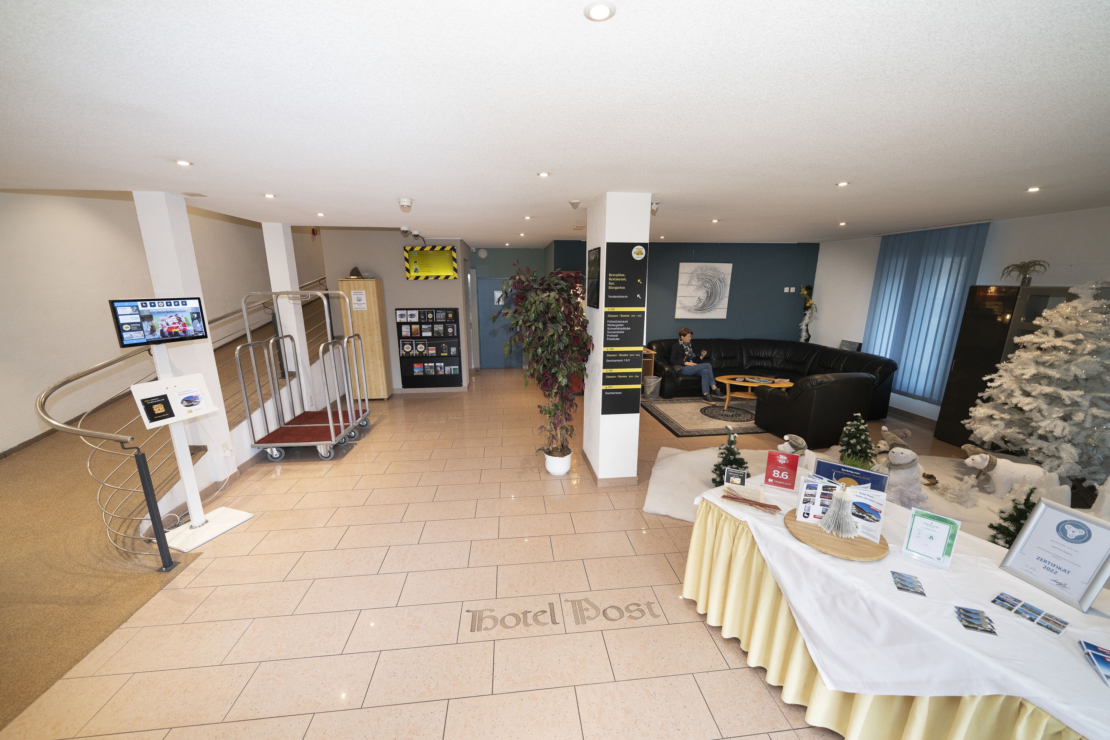 A hotel lobby with a reception desk, a staircase, a seating area, and a display table with brochures.