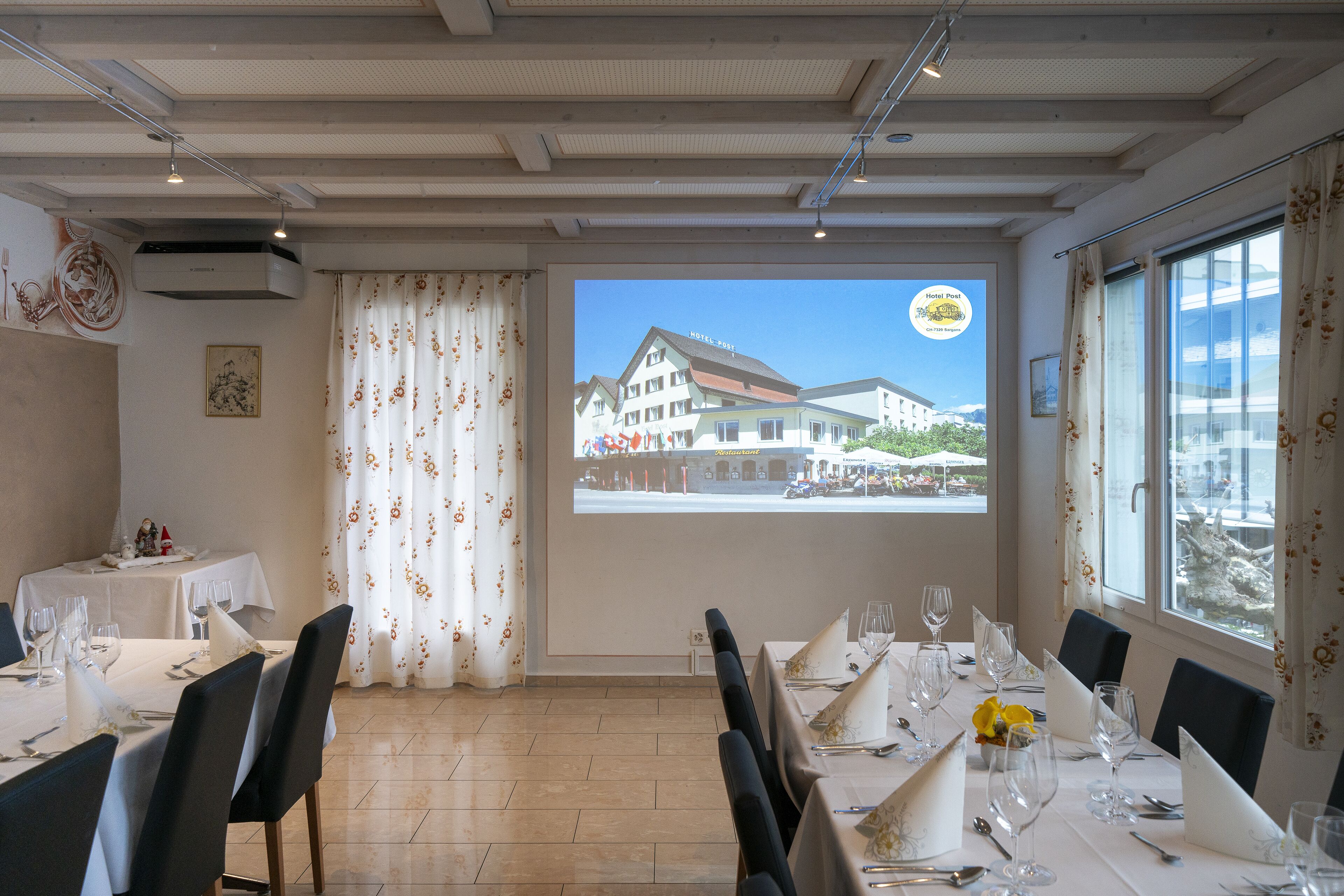 Ein Essbereich mit gedeckten Tischen, einem großen Fenster mit Vorhängen und Blick auf ein Hotelgebäude.