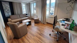 A modern living room with a sofa, coffee table, and a desk with a chair. There are large windows with curtains, a radiator, and a view of buildings outside. - Hotel Post (Sargans)