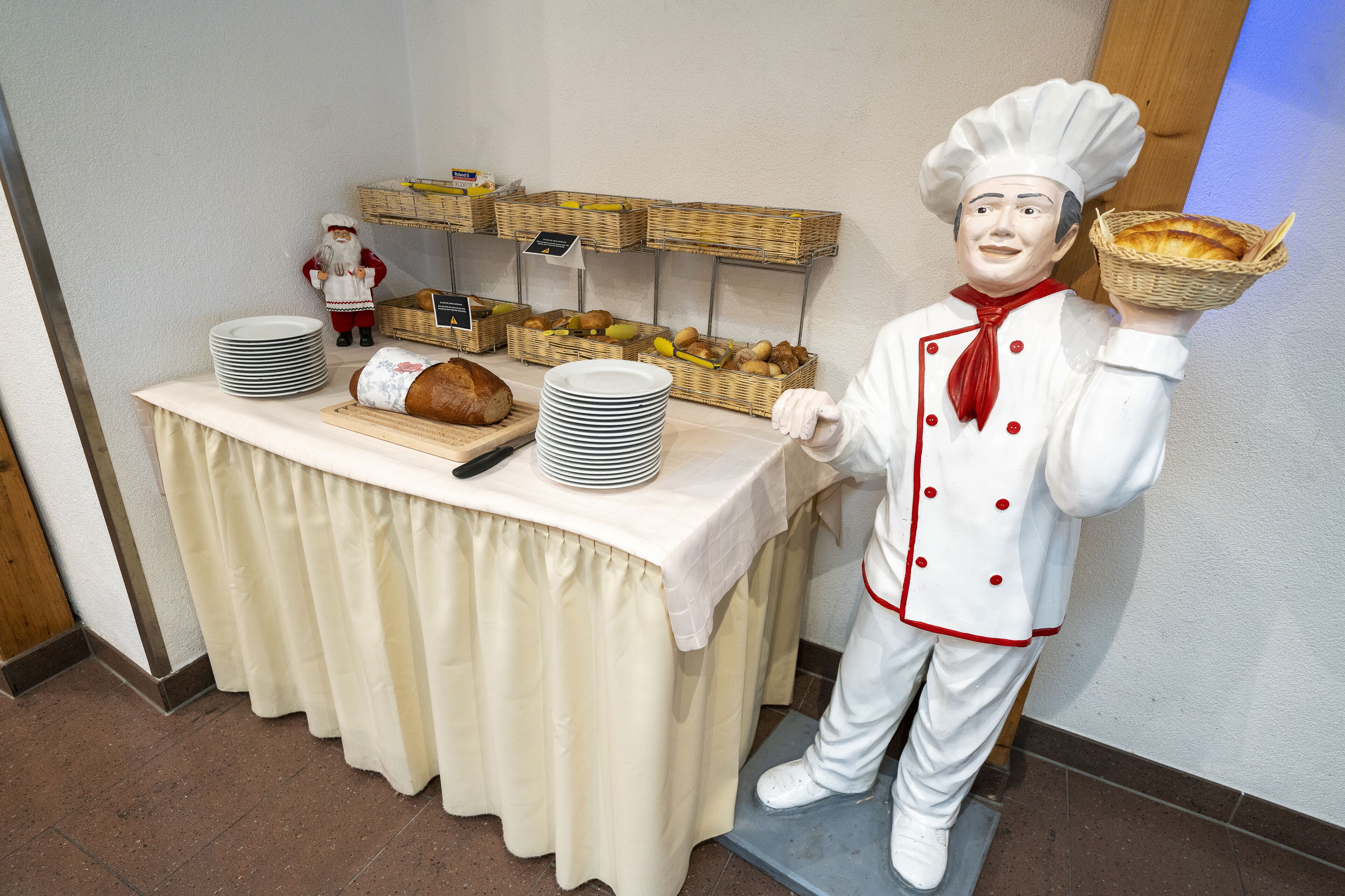 A buffet setup with a chef statue holding a basket of bread, plates stacked on a table, and baskets of fruits and bread on shelves.