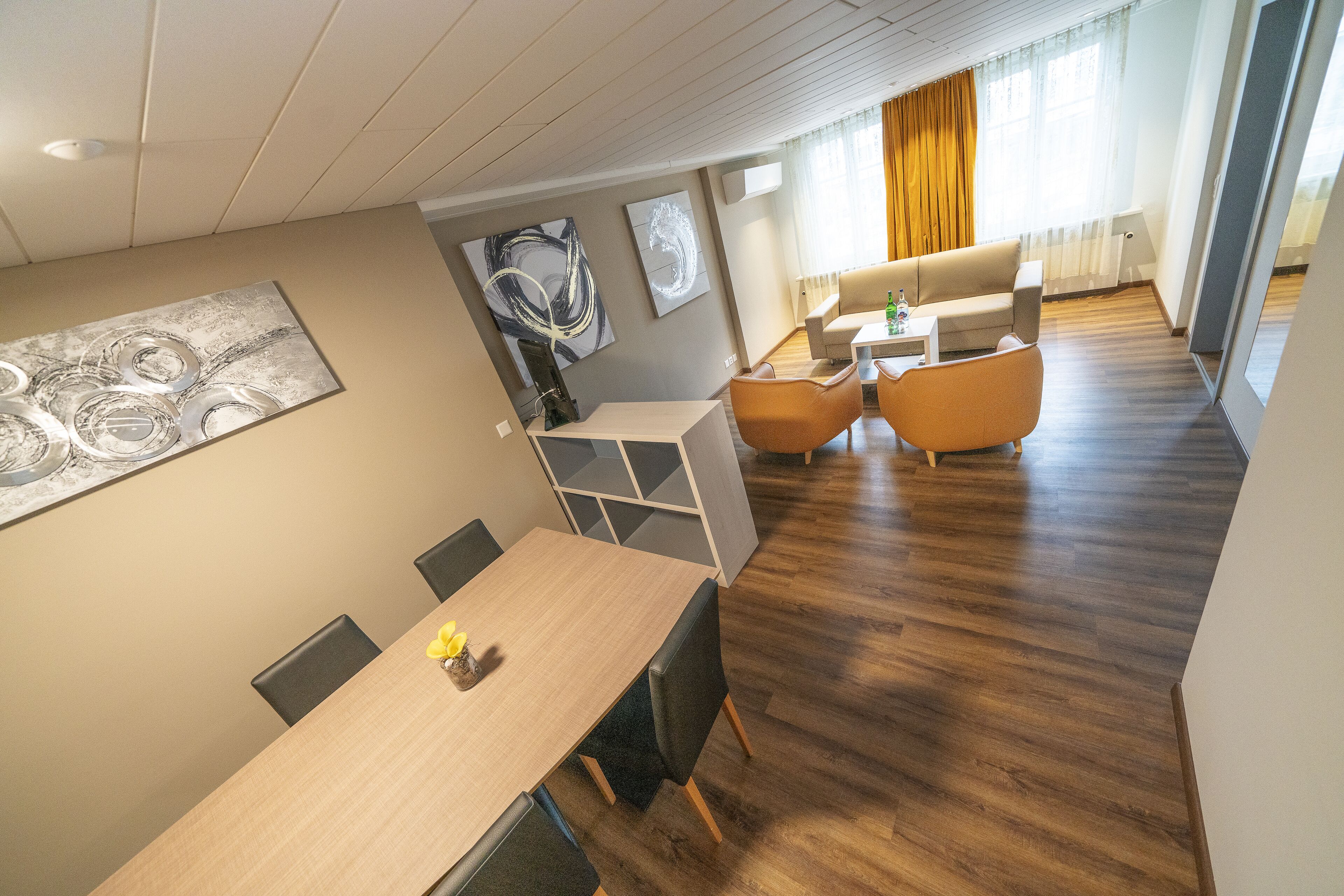 Una habitación de hotel moderna con una mesa de comedor de madera, sillas, un sofá y una mesa de centro. Se observan obras de arte abstracto en las paredes y una ventana grande con cortinas.