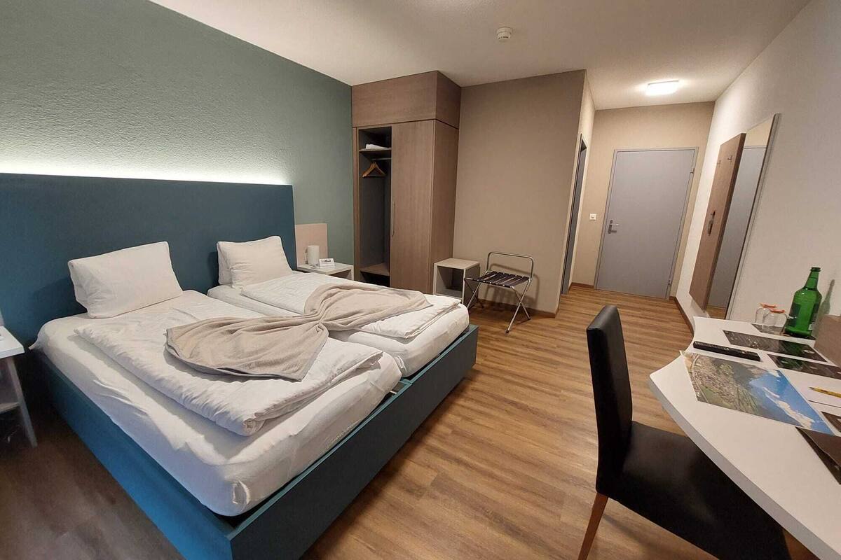 A modern hotel room with a large bed, a desk with a chair, a small table, and a wardrobe.
