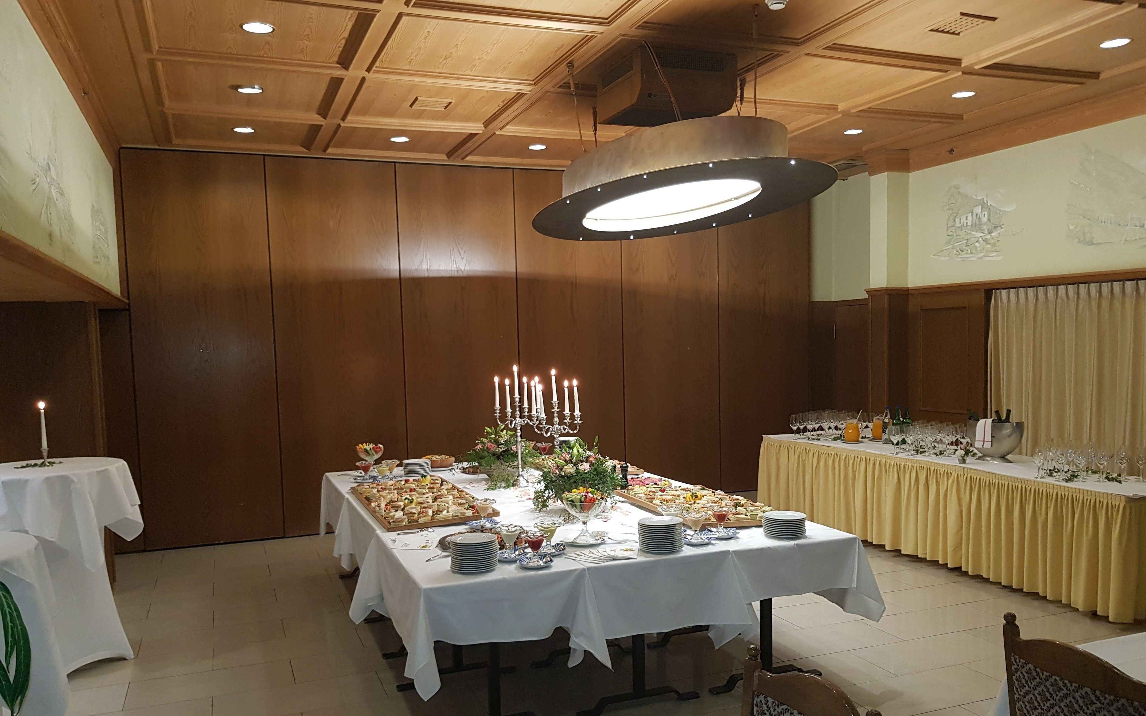 A banquet hall with a long table set for a buffet, featuring various food items and a centerpiece with candles.