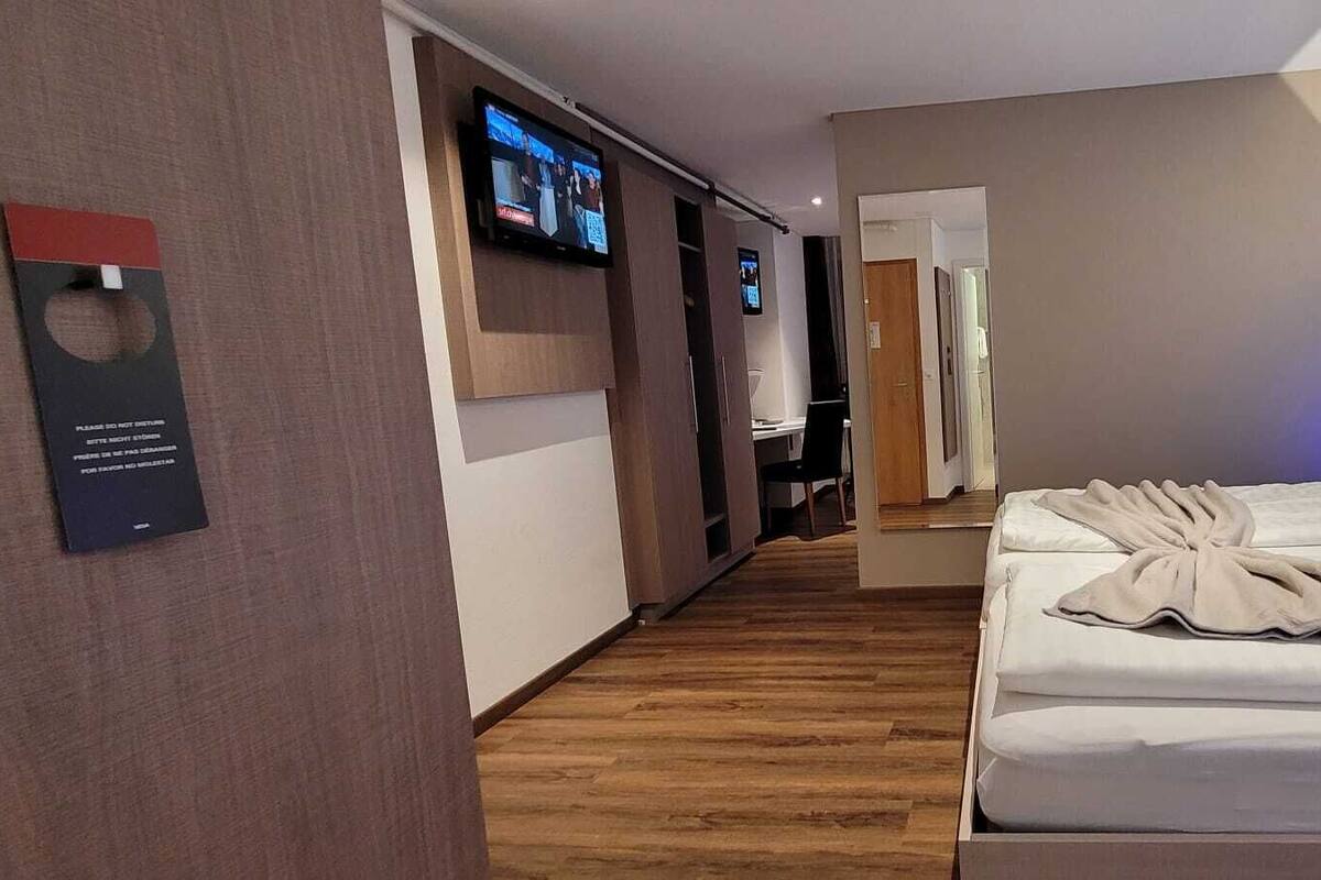 A hotel room with a wooden floor, a bed with a white comforter, a TV mounted on the wall, and a small desk with a chair.