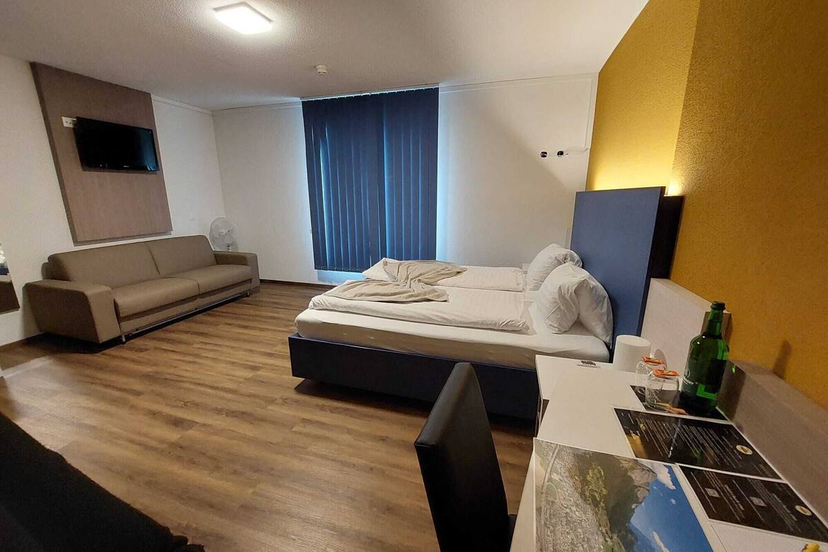 A modern hotel room with a bed, a sofa, a flat-screen TV mounted on the wall, and a large window with curtains.
