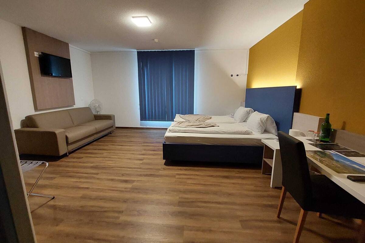 A hotel room with a bed, a desk, a chair, a sofa, a fan, and a window with curtains.