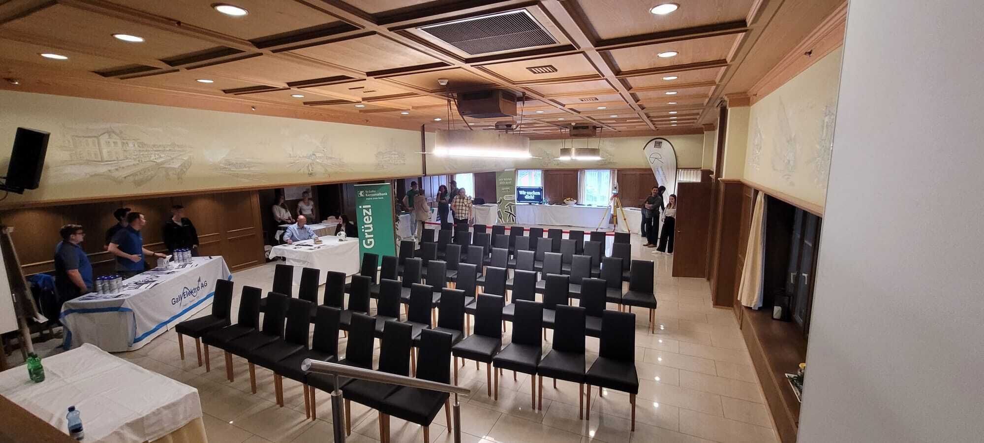 A conference room with rows of chairs, a table with a banner, and a stage area.