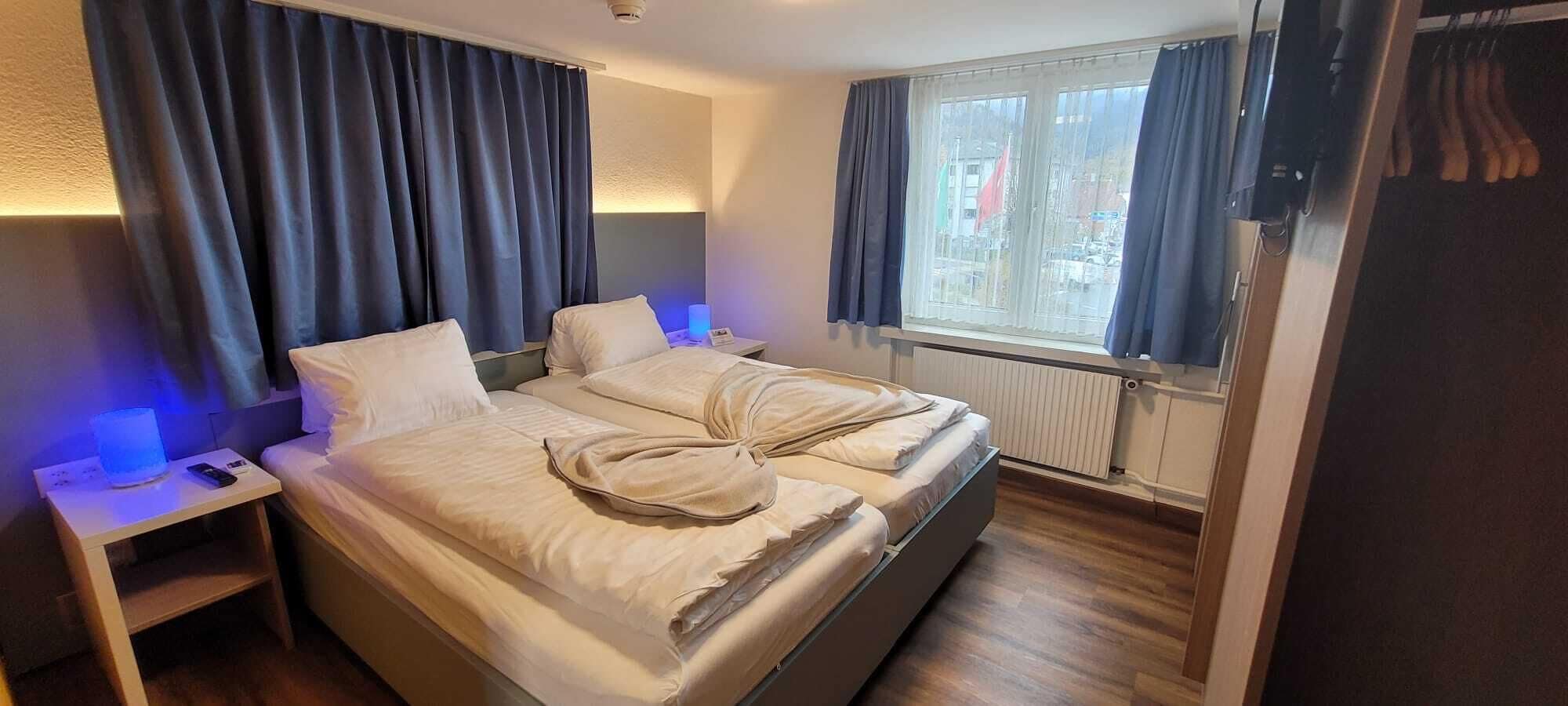A double bed with a blanket, a bedside table with a lamp, and a window with curtains.